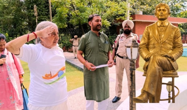 A gilded statue of Madan Lal Dhingra seated in a chair stands in a memorial park in Amritsar; visitors and a uniformed police officer stand nearby, one of them saluting.