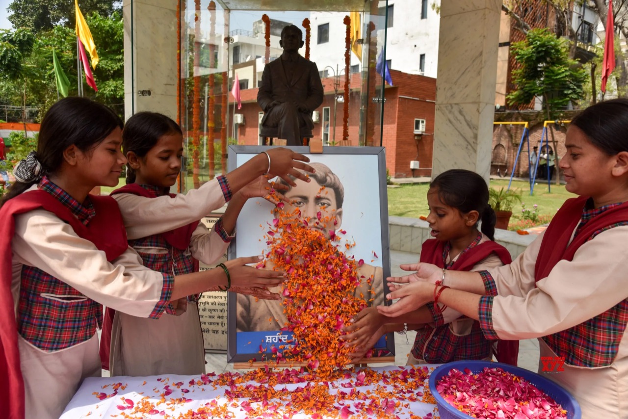Four schoolgirls in uniform scatter marigold and rose petals over a framed portrait of Madan Lal Dhingra at an outdoor memorial; behind the frame, a bronze seated statue of him stands inside a glass pavilion hung with flower garlands.