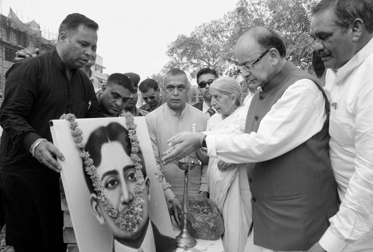 A black and white photograph of a commemorative ceremony: a group of people stand around a garlanded framed portrait of Madan Lal Dhingra while one of them lights a tall brass lamp.