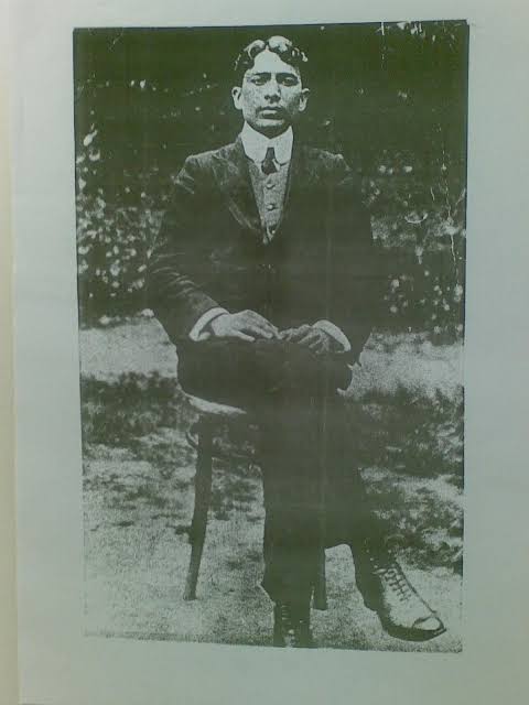 Full-length photograph of Madan Lal Dhingra as a young man, seated on a bentwood chair in a garden, wearing a dark three-piece suit and boots, hands resting in his lap.